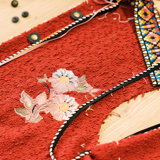 Roter Trachtenstoff mit floralem Stickmotiv, schwarzer Borte und buntem Webband liegt zugeschnitten mit sichtbaren Stoffkanten auf einem Holztisch image number