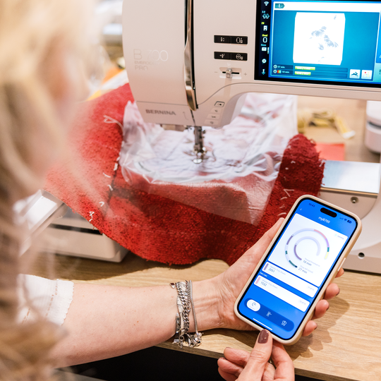 Person bedient eine BERNINA 700 PRO Stickmaschine, w&auml;hrend sie auf einem Smartphone eine verkn&uuml;pfte Stick-App mit Einstellungsanzeige nutzt. image number